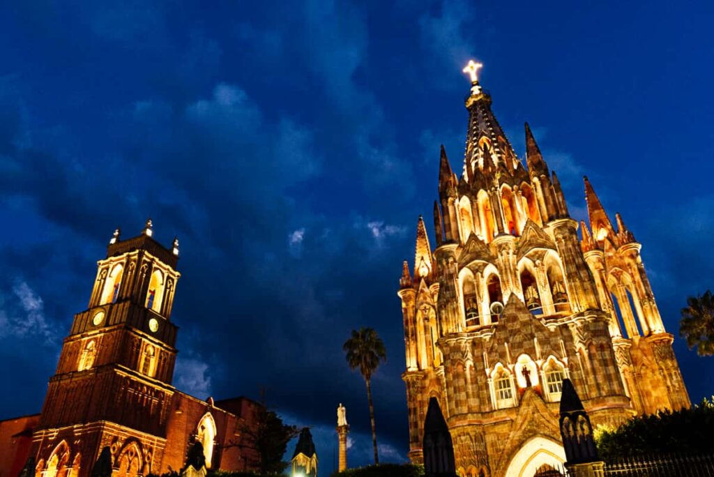 The main church in San Miguel de Allende, with all of its architecture lit up at night. On the very top is a lit cross. Dark blue sky behind and palm trees on either side.