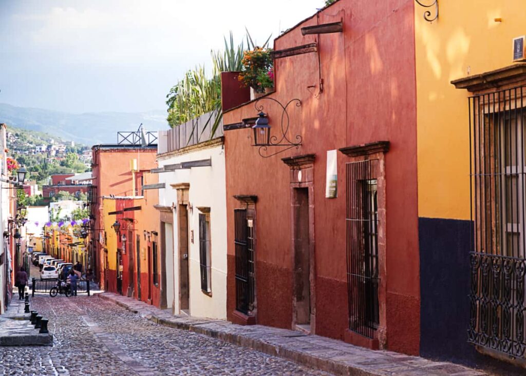 Buildings in hues of red and ochre line the streets, one of several sights on a San Miguel de Allende tour. In the background are more buildings and low mountains.