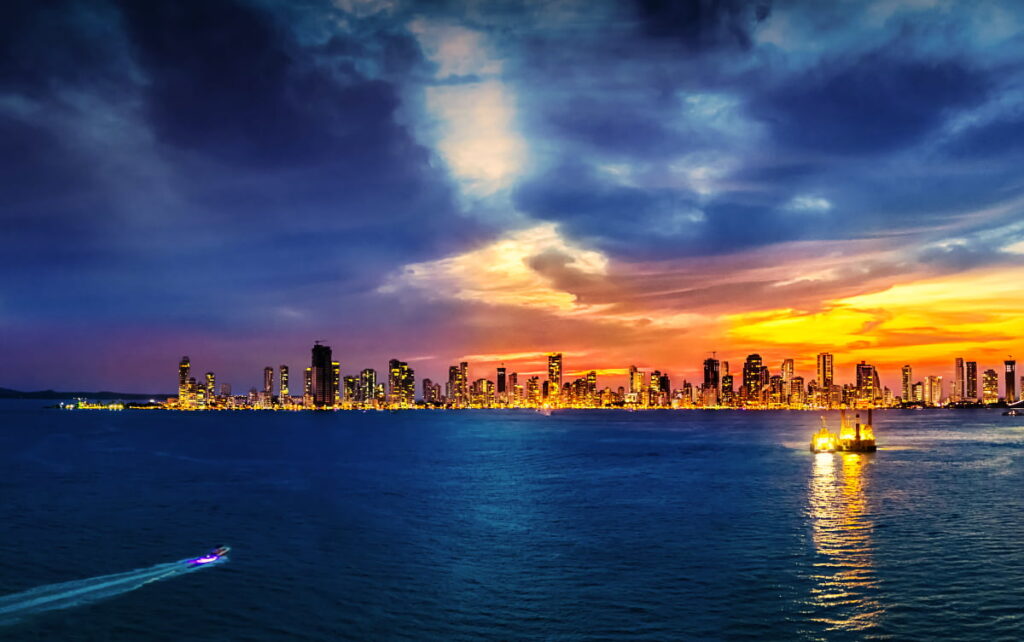 On this sunset boat tour, Cartagena skyline is lit up with lights from the buildings. Above, the sky is hues of orange, pink, and purple from the sunset.