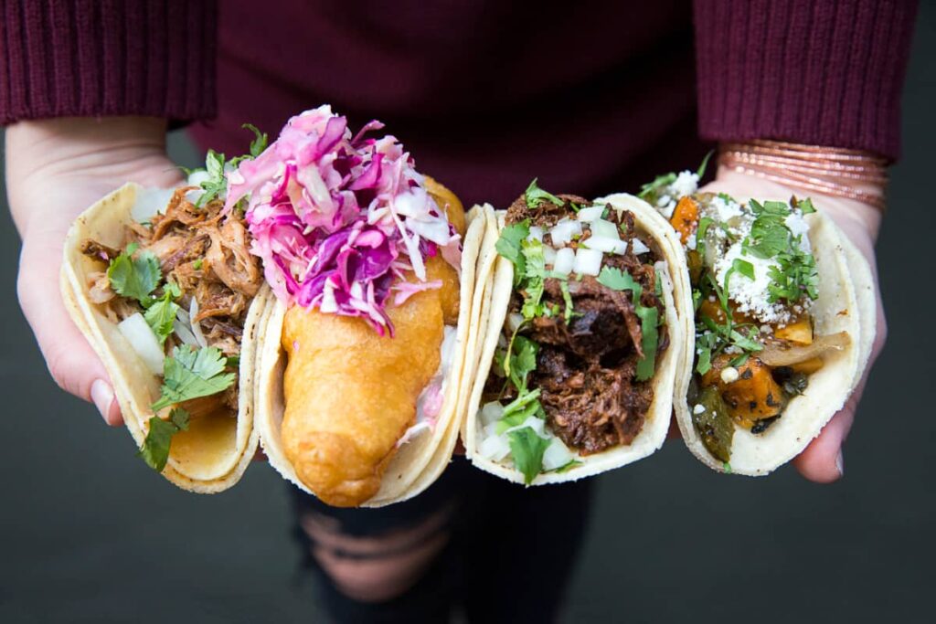 A woman holds four different types of tacos during a taco tour in Mexico City. They include Baja style seafood topped with purple cabbage, carnitas tacos, barbacoa, and a vegetarian taco.