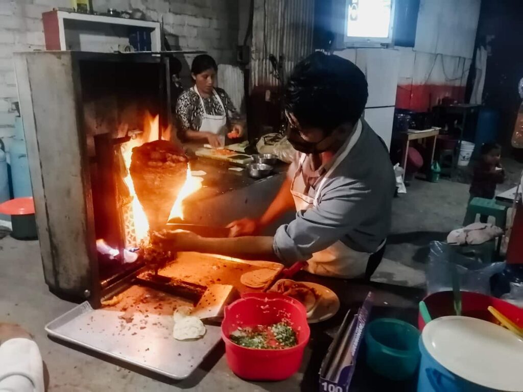 During a Mexico City taco tour, a man cuts meat from a spit rotating in front of a flame. In the background a woman assembles tacos al pastor.