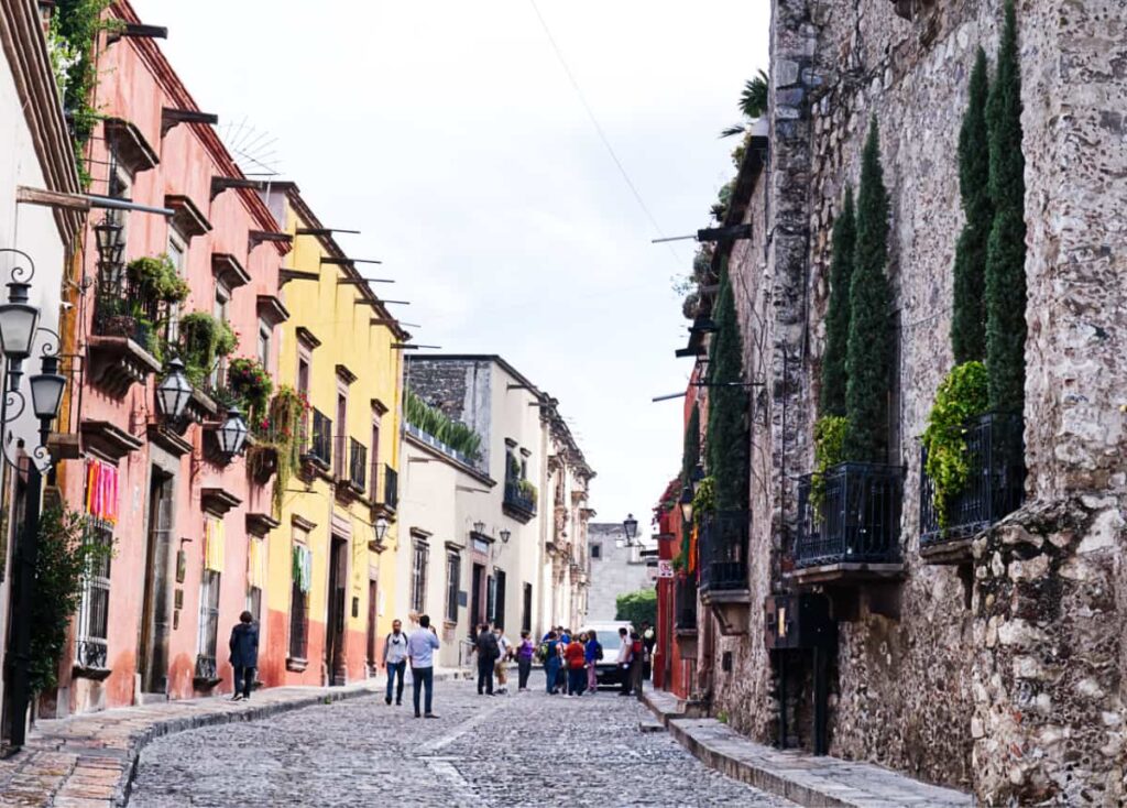 During a walking tour in San Miguel de Allende, this street showcases buildings in a bright pink and yellow. People are gathered on the cobblestone street which is also lined with a grey stone building on the right.