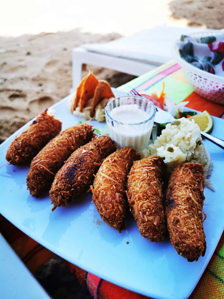 Six pan fried coconut shrimp lined up on a plate, one of the most popular foods to eat on Ixtapa Island. Also on the plate are small portions of rice, salad, and tortilla chips as well as a shot glass of coconut cream. The sandy beach is seen in the background.