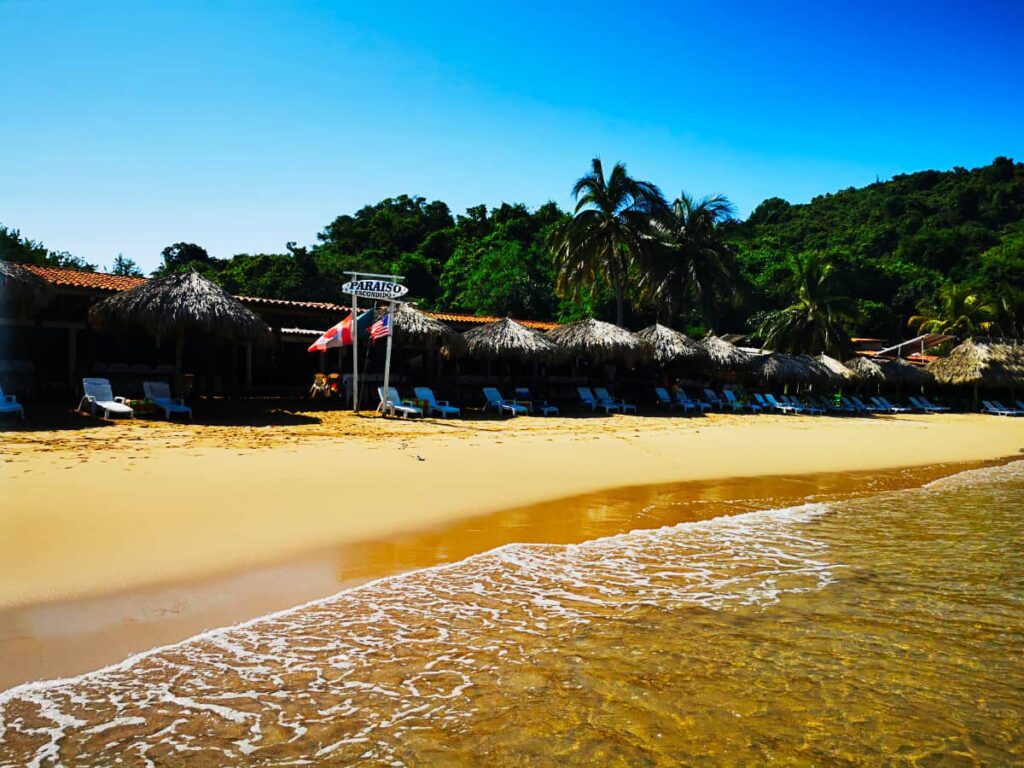 A shallow waves comes on shore in front of one of the Ixtapa Island restaurants whose surfboard shaped sign says Paraiso Escondido (Hidden Paradise). Along the beach are several lounge chairs shaded under the palapas.