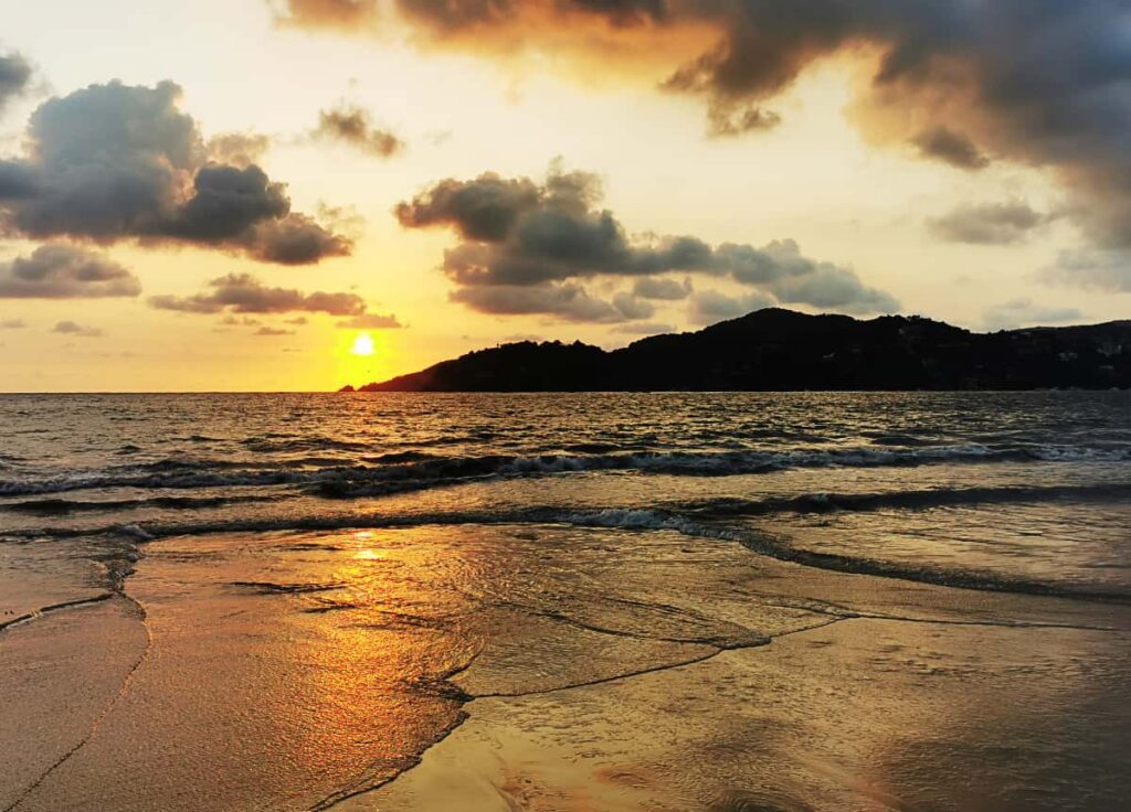 During sunset, light waves come to shore on La Ropa Beach Zihuatanejo. The setting sun casts a golden tone onto the clouds and beach while silhouetting the mountain in the background.