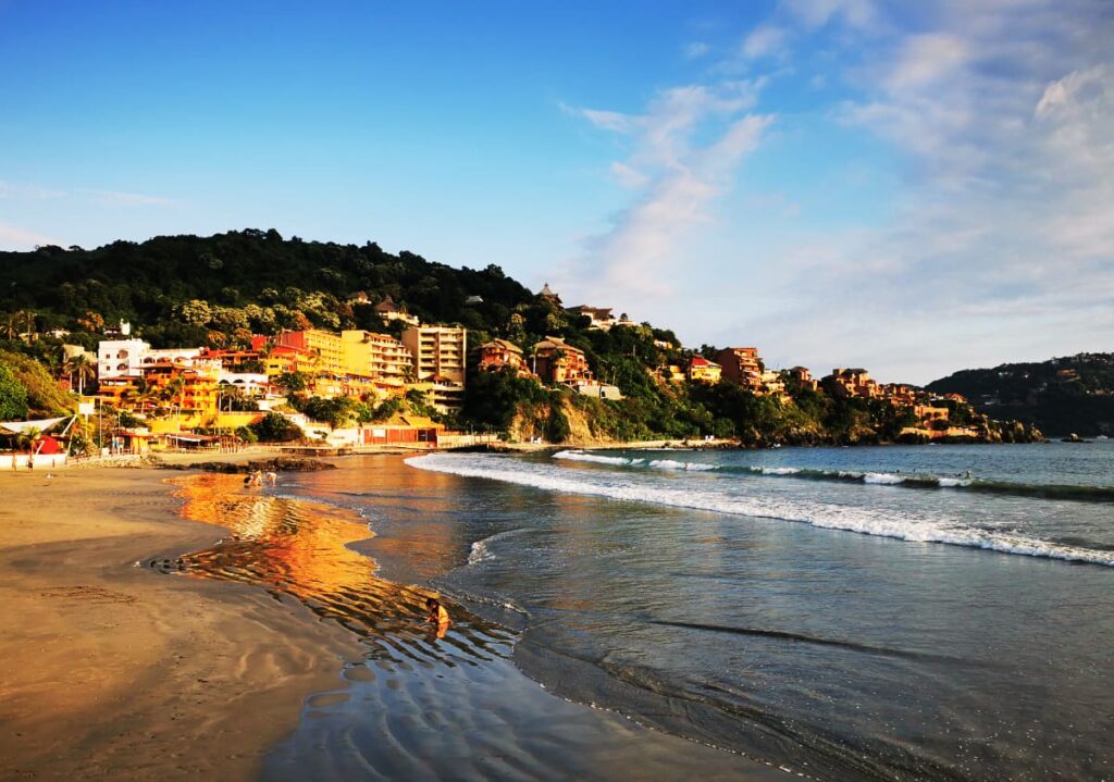 A child plays in the surf at Madera Beach in Zihuatanejo as shallow waves come to shore. Behind the hillside and buildings are lit by the golden sun which is also reflected in the wet sand below.