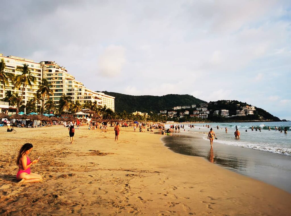 A woman in a pink bikini sits in the sand at Playa Palmar, the main beach in Ixtapa Zihuatanejo. Other people swim in the ocean, walk along the waves, and play on the beach. In the background are several Ixtapa resorts.