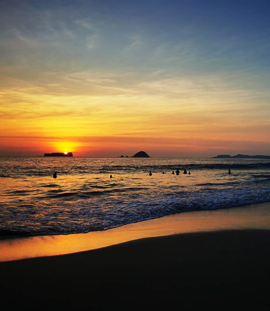 Several people play in the waves during sunset on Playa el Palmar in Ixtapa Zihuatanejo. The golden orange hues reflect into the sky and the ocean below.