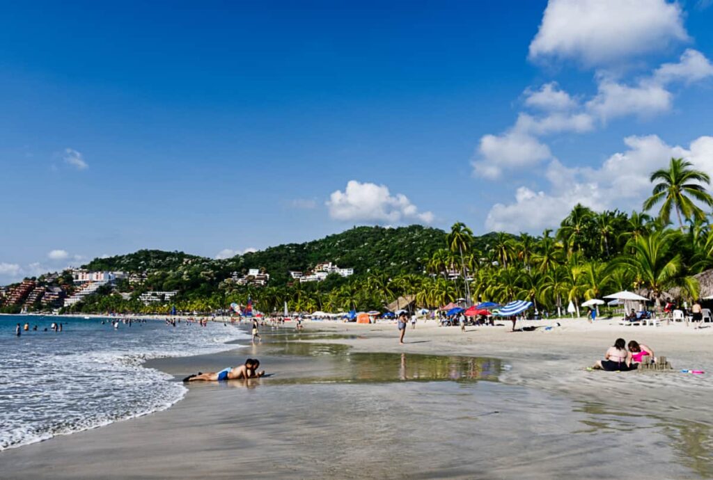 People play and lounge in the sand lined with palm trees at Playa Ropa in Zihuatanejo. In the background are several hotels and resorts.