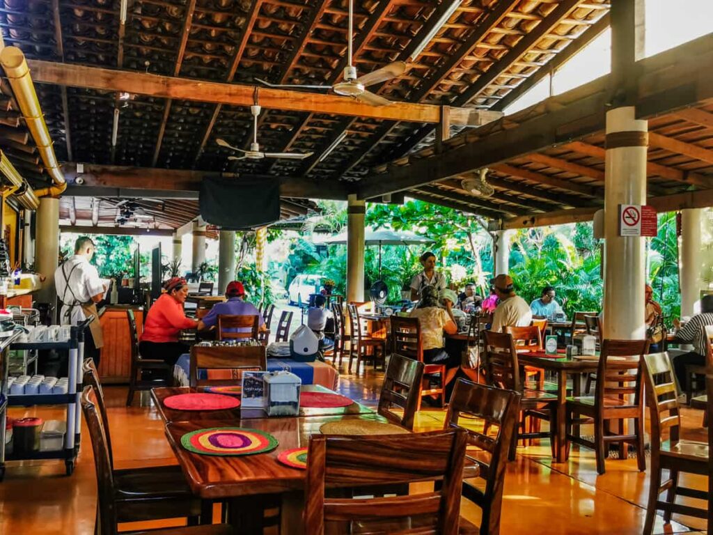 A patio setting surrounded by tropical plants in a Zihuatanejo breakfast restaurant, with wooden furniture and colorful table settings,.