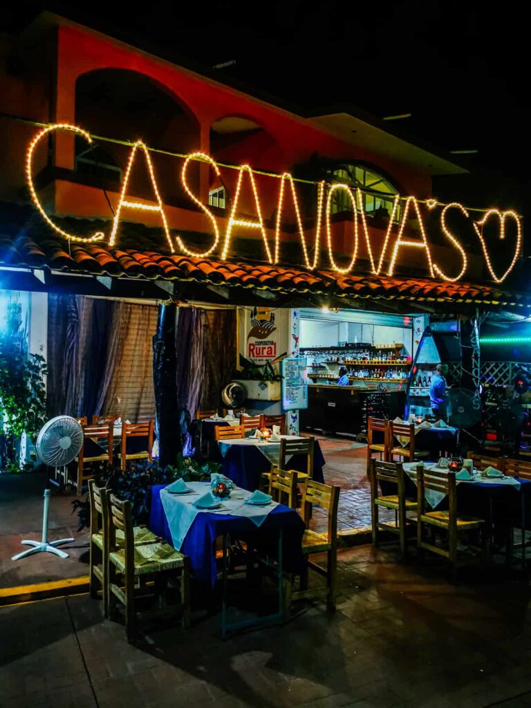 The glowing sign of 'Casanovas' stands out against the evening sky at this top restaurant in Zihuatanejo. Several tables with tablecloths are outside, in front of a fan.