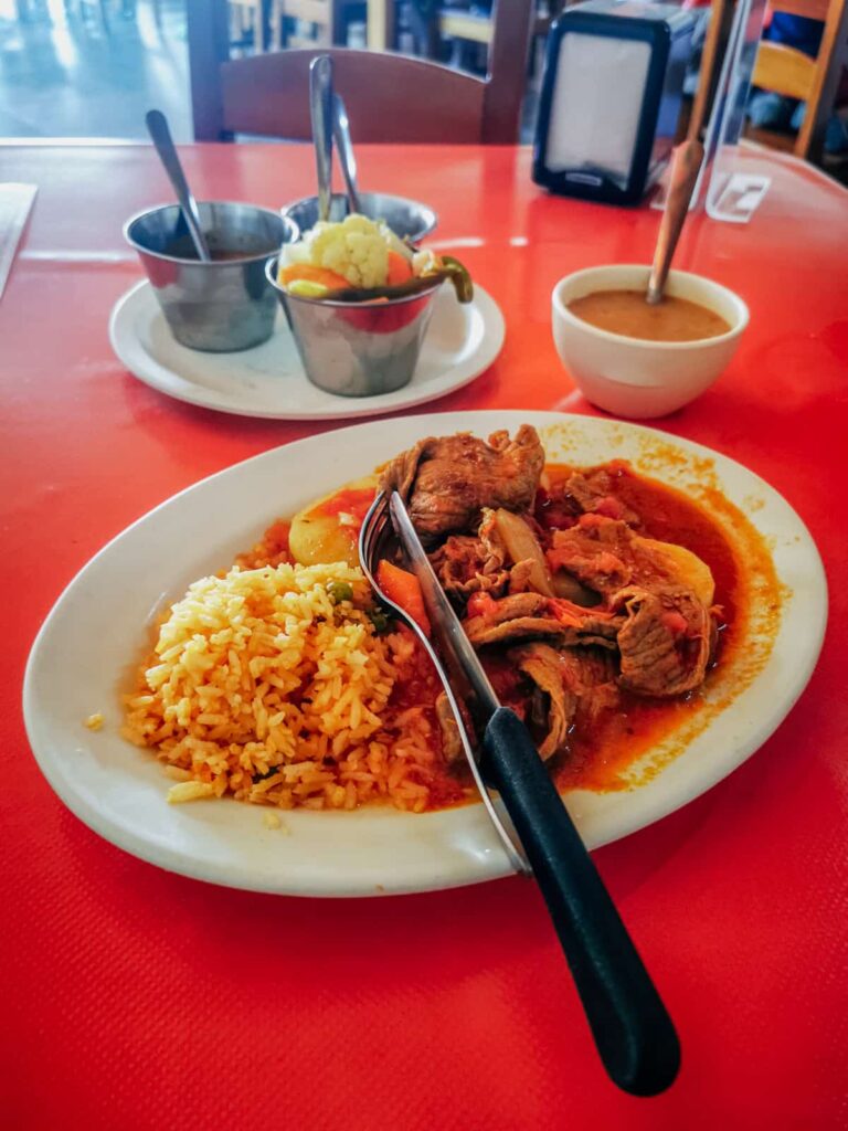 A plate of traditional Mexican stew served with rice and vegetables, presented on a white plate against the bright red table of an affordable place to eat lunch in Zihuatanejo.