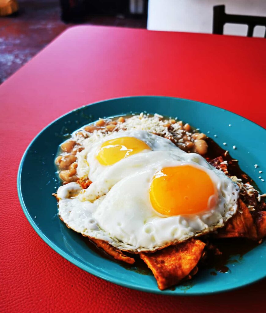 A plate of traditional Mexican chilaquiles served at a restaurant in Ixtapa, featuring crispy tortillas smothered in red sauce, topped with two sunny-side-up eggs, and accompanied by refried beans, all on a vibrant turquoise plate against a red table.