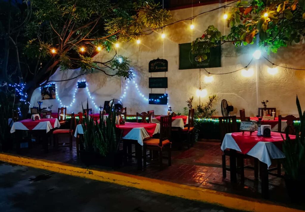 An inviting outdoor seating area at El Arrayán restaurant in Zihuatanejo, with string lights twinkling among the trees and plants, creating a cozy atmosphere. The tables are set with red and white tablecloths, ready for diners, against the backdrop of a warmly lit white wall adorned with a TripAdvisor certificate of excellence banner.