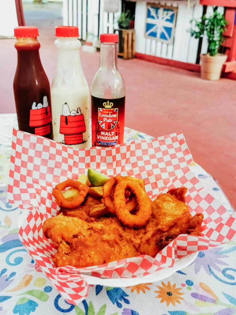 A classic plate of fish and chips in Zihuatanejo are served in a red and white checkered basket, with onion rings and a side of lime. On the table are bottles of tartar sauce and malt vinegar.