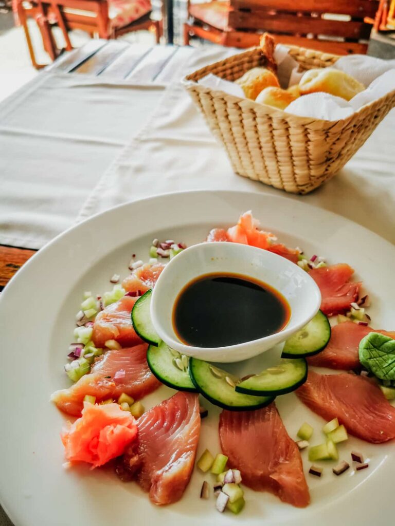 A plate of fresh tuna sashimi, elegantly arranged with slices of cucumber and diced red onion, served with soy sauce and a garnish of ginger, presented on a white plate with a basket of bread rolls in the background at a fine dining restaurant in Zihuatanejo