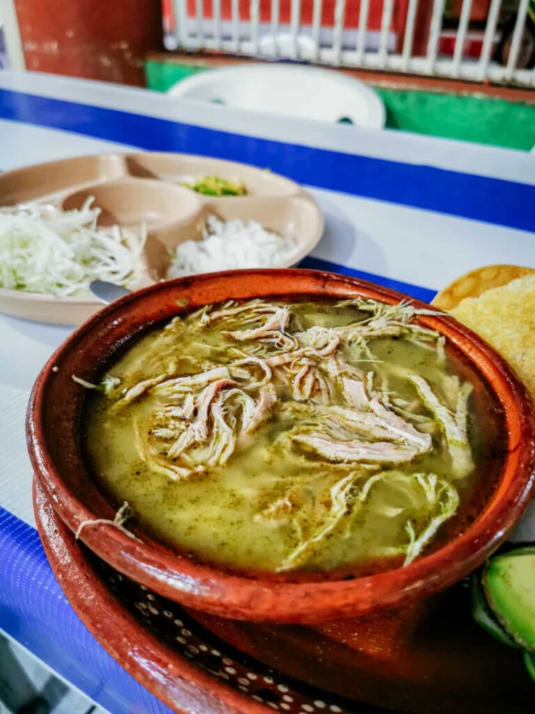 A soup bowl of green pozole, filled with shredded pork and is served alongside shredded cabbage, diced onion, and tostadas, and chicharron in a restaurant in Zihuatanejo, Guerrero.