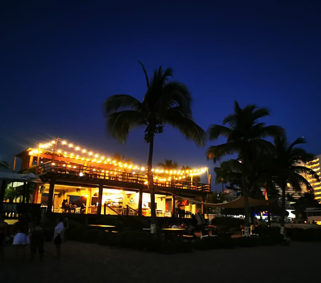 A picturesque beachfront restaurant in Ixtapa at dusk, illuminated by string lights against the twilight sky. Palm trees frame the scene, adding tropical ambiance as patrons enjoy dining with a view of the ocean.