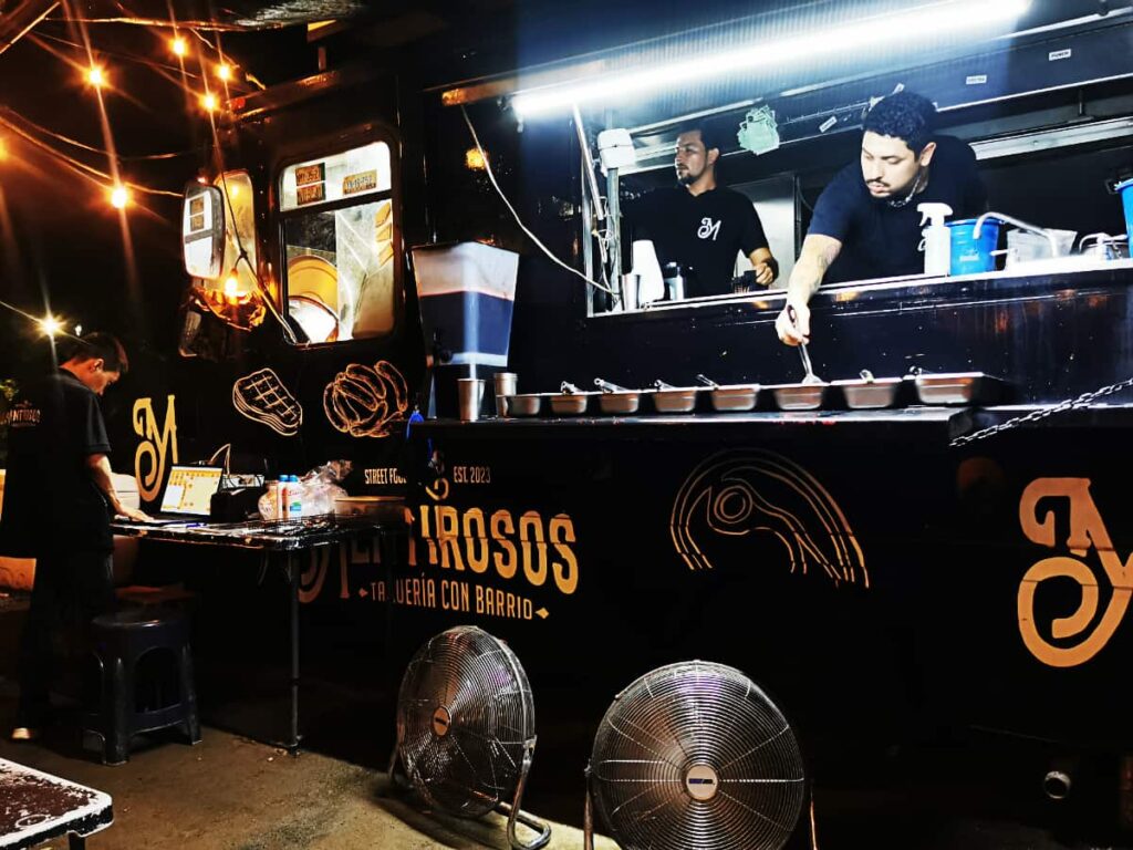 A bustling taco truck glows under string lights at night, with chefs busily preparing food. The truck's dark color with vibrant signage and the presence of fans for customer comfort create an inviting place to eat street food in Ixtapa.