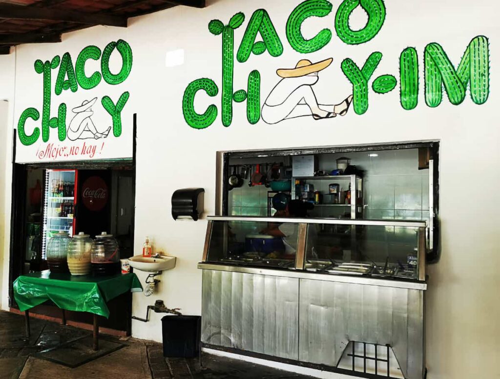 A casual restaurant to eat tacos in Ixtapa, featuring a prominent green "TACO CHAY" sign with the figure of a man in a sombrero used to create the letter A. The counter displays a clear view into the kitchen, and beverage dispensers are ready for service, inviting patrons to enjoy a authentic Mexican tacos.