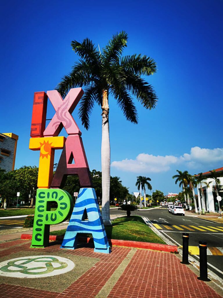 Colorful letters are stacked vertically spelling out Ixtapa. Behind is a palm tree and the main street in Ixtapa Zihuatanejo.
