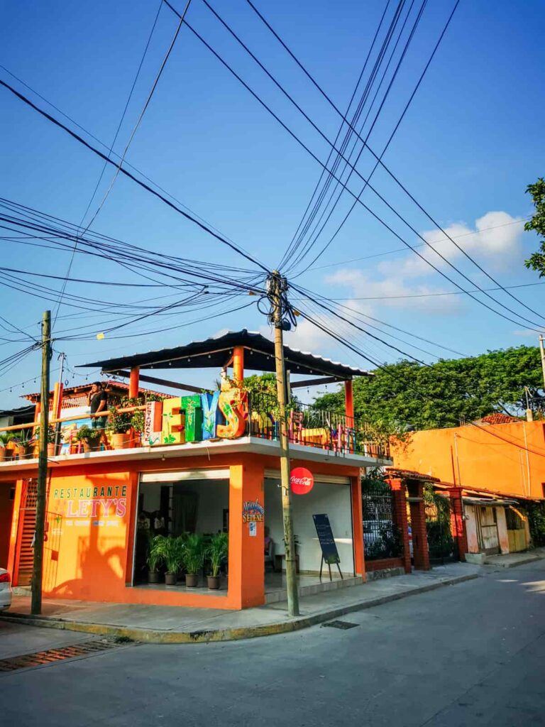Exterior view of Lety's Restaurant in Zihuatanejo, Mexico, showcasing its vibrant orange facade and second-story dining terrace adorned with colorful banners. The corner setting is accentuated by an intricate web of power lines against a clear blue sky.