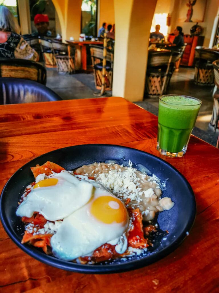 A traditional Mexican breakfast of chilaquiles topped with two sunny-side-up eggs and crumbled cheese, accompanied by a side of beans, served with a refreshing glass of green juice, in a cozy Zihuatanejo restaurant.