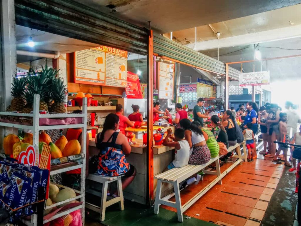 Vibrant local dining scene at Zihuatanejo market with families enjoying traditional breakfast at a restaurant in the market surrounded by fresh fruit displays.