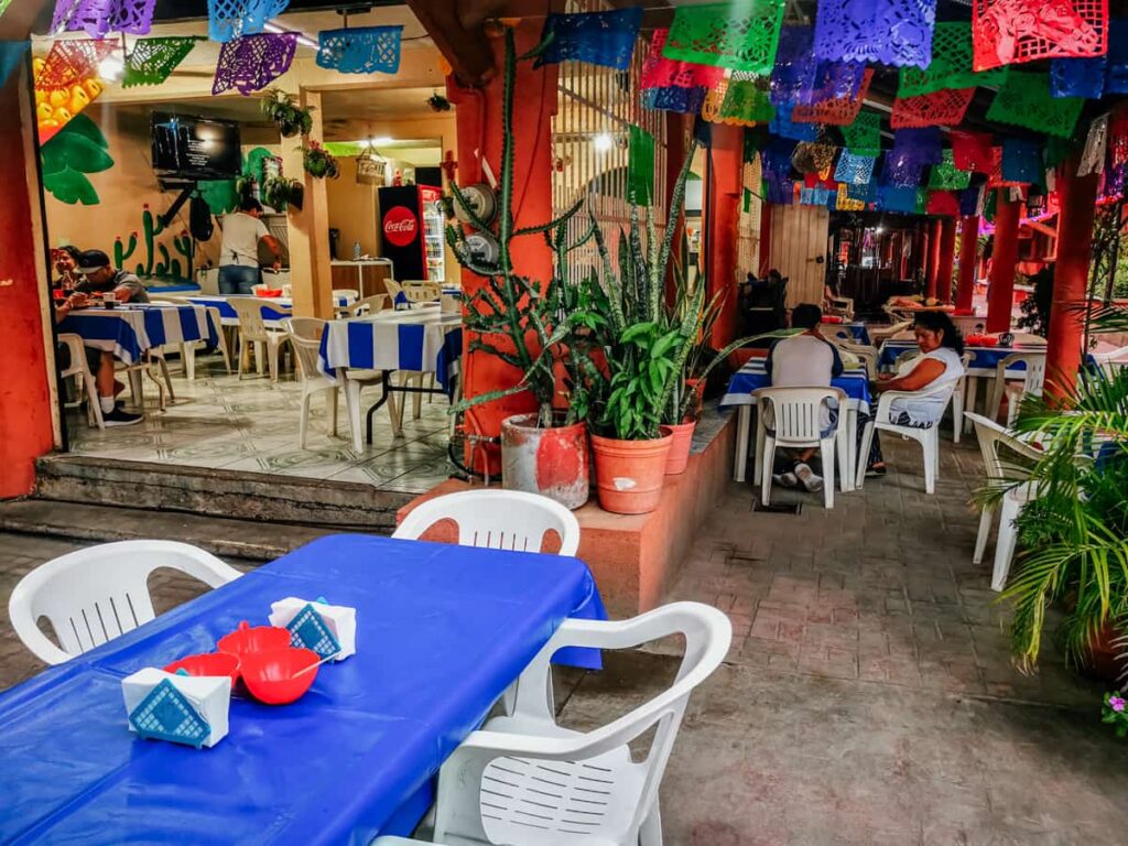 An outdoor restaurant in Zihuatanejo with colorful papel picado streamers, blue and white striped tablecloths, and potted plants creating a welcoming atmosphere for casual dining.