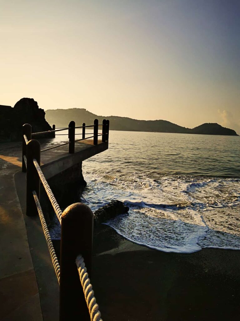 Sunrise view of Paseo del Pescador in Zihuatanejo, with gentle waves lapping against the pier and golden sunlight silhouetting the tranquil bay. The elevated paved walkway is lined with a rope railing.