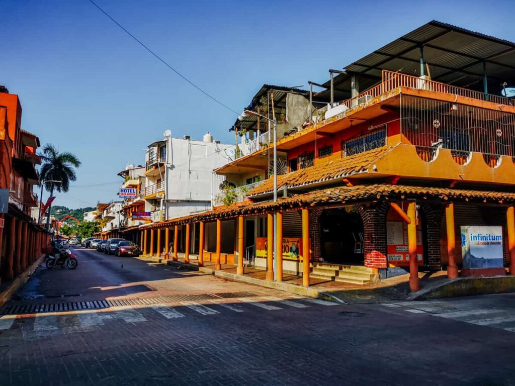 A serene scene shows a sunlit street in downtown Zihuatanejo, flanked by orange-hued buildings with terracotta roofs over the sidewalks under a clear blue sky.