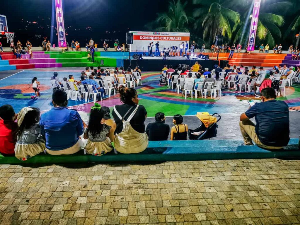 A family sits on the edge of Zihuatanejo's basketball court for a Sunday evening cultural event. In front of them the stage advertises "Domingos Culturales" and people are sitting in white plastic chairs in front of the stage.