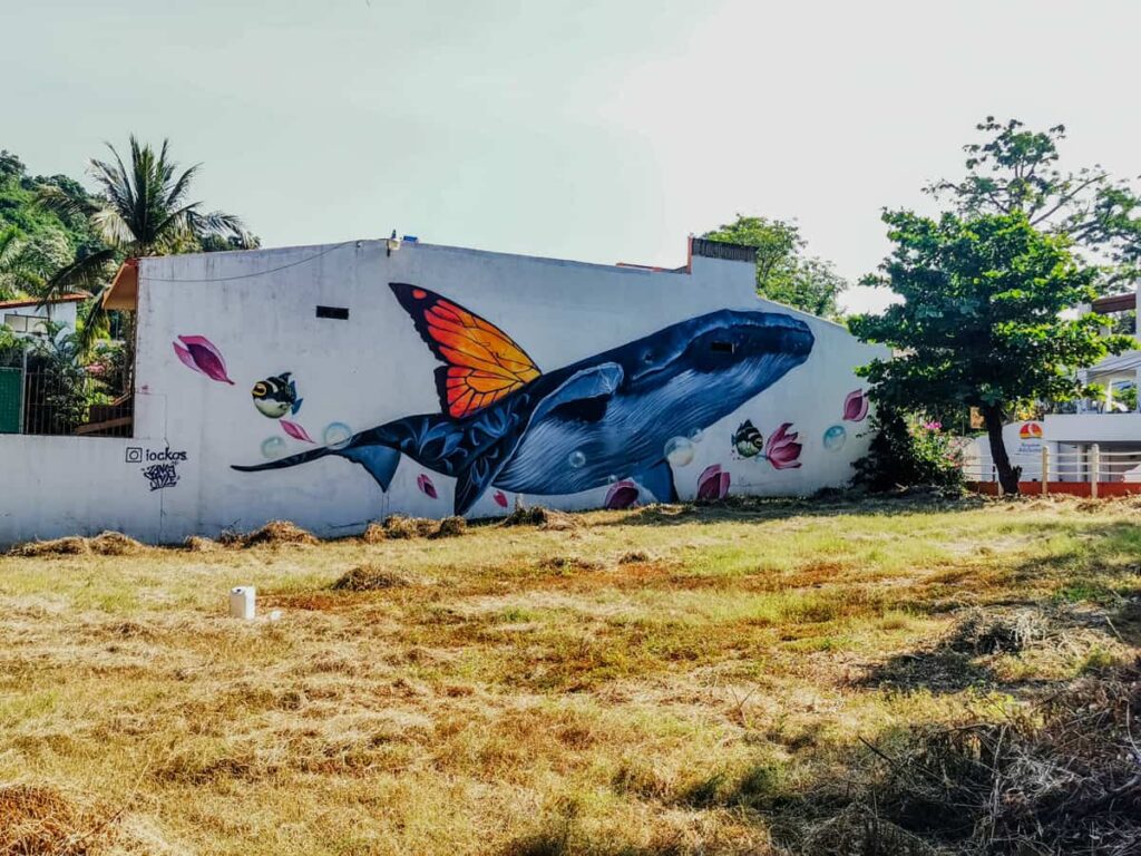 Large street art on the side of a building in La Madera Zihuatanejo depicts a blue whale with monarch butterfly wings, fish, and bubbles.