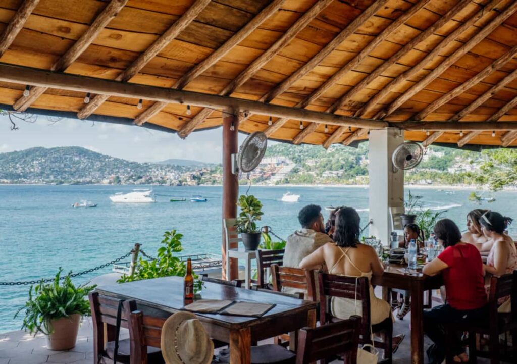 Patrons dine at a cozy, open-air Zihuatanejo restaurant with a view of the bay, where boats are anchored in the water. The setting is rustic with wooden furniture and a wooden roof, accented with potted plants.