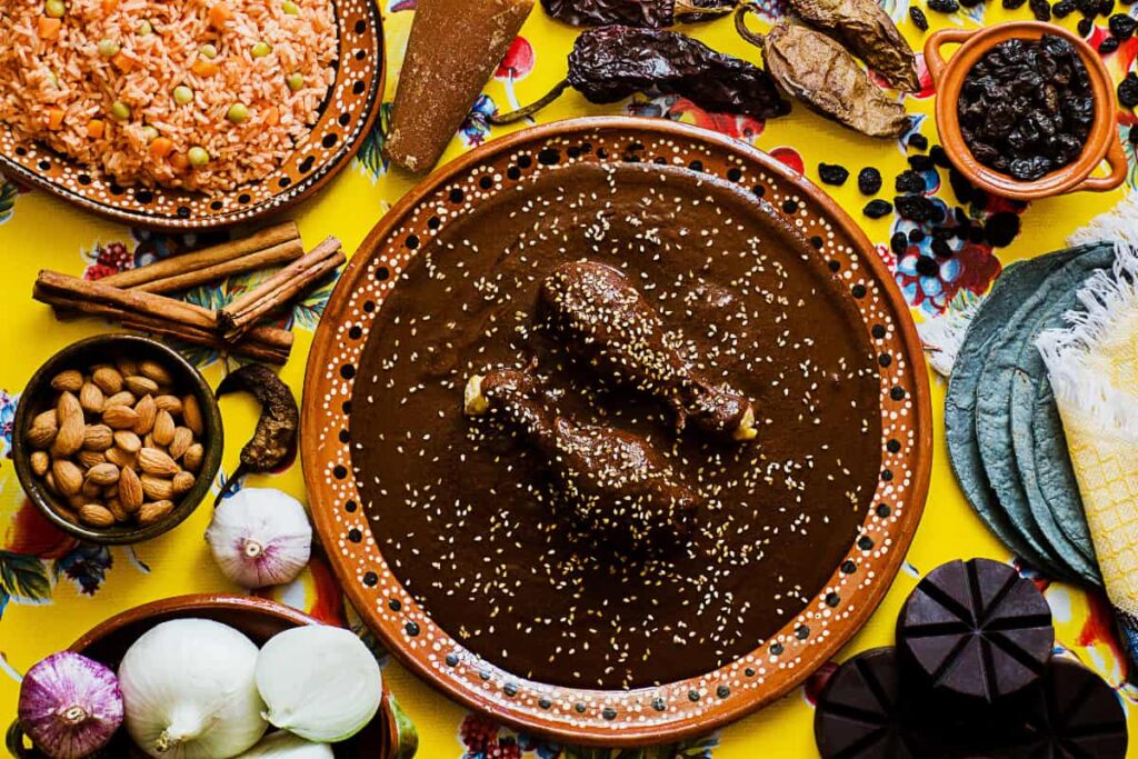 An overhead view of a table at a Mexico City cooking class includes a clay plate of two chicken legs covered in a brown mole sauce topped with sesame seeds. Around the plate are the ingredients to cook mole, tortillas, and a bowl of red rice with peas.