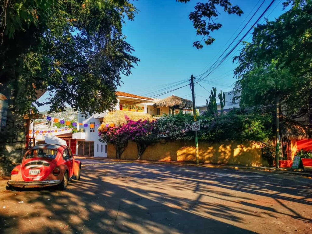A quiet street scene in La Madera Zihuatanejo Mexico depicts a bright orange VW Beatle parked on the side of the shady street.