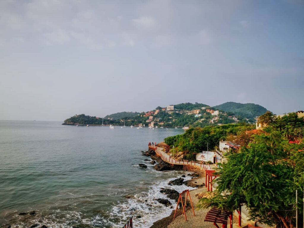 Scenic view of the bay in Zihuatanejo Mexico with its calm waters, rocky shoreline, and lush hillsides dotted with buildings. The walkway, paseo del pescador, snakes along the oceanfront below.