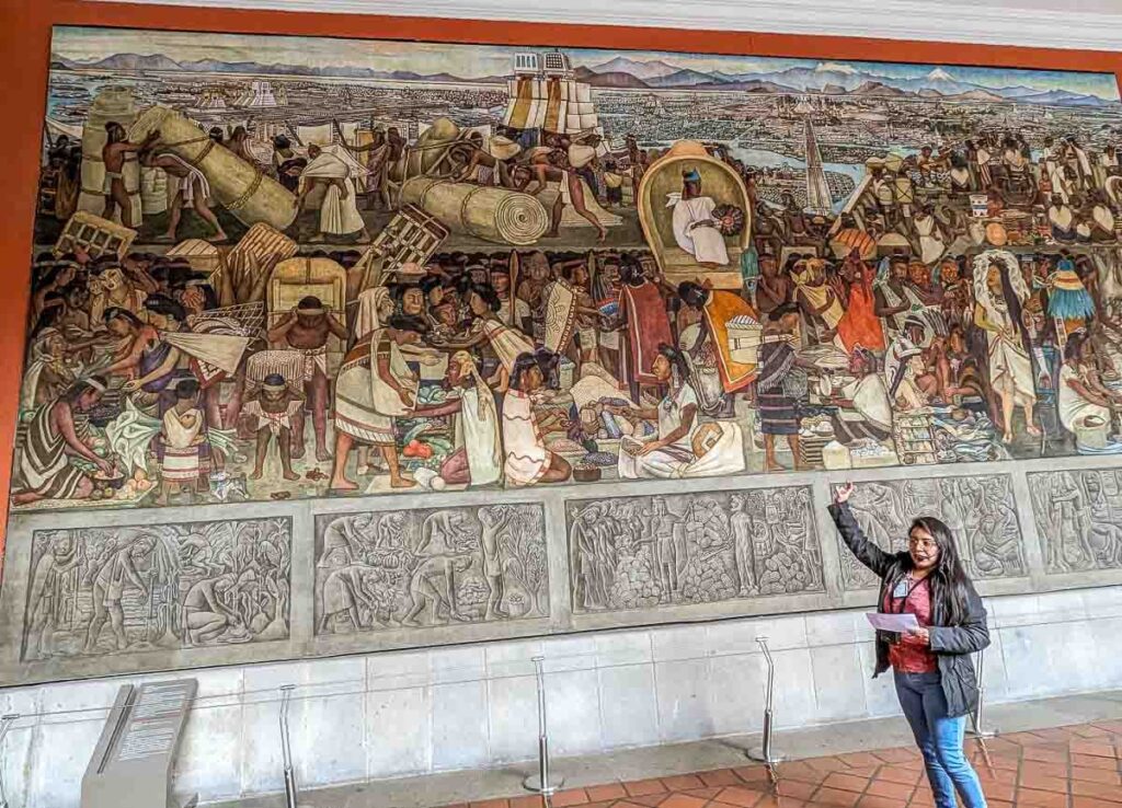 A tour guide pointing to a Diego Rivera mural depicting ancient Mexico during a National Palace tour in CDMX.