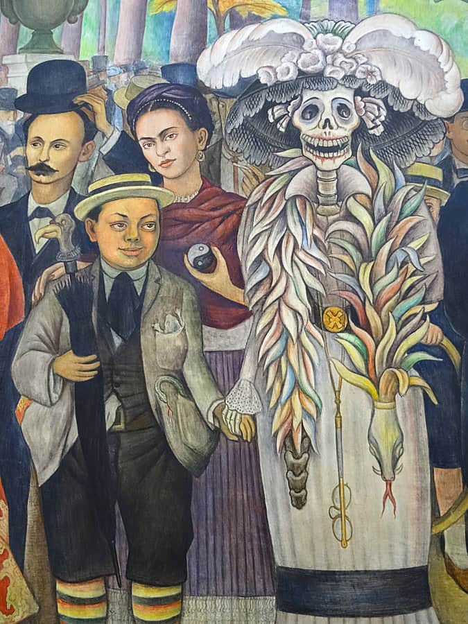Detail of a Diego Rivera mural that depicts the artists as a young boy holding the hand of a skeleton Catrina figure with Frida Kahlo behind him holding a yin yang symbol.