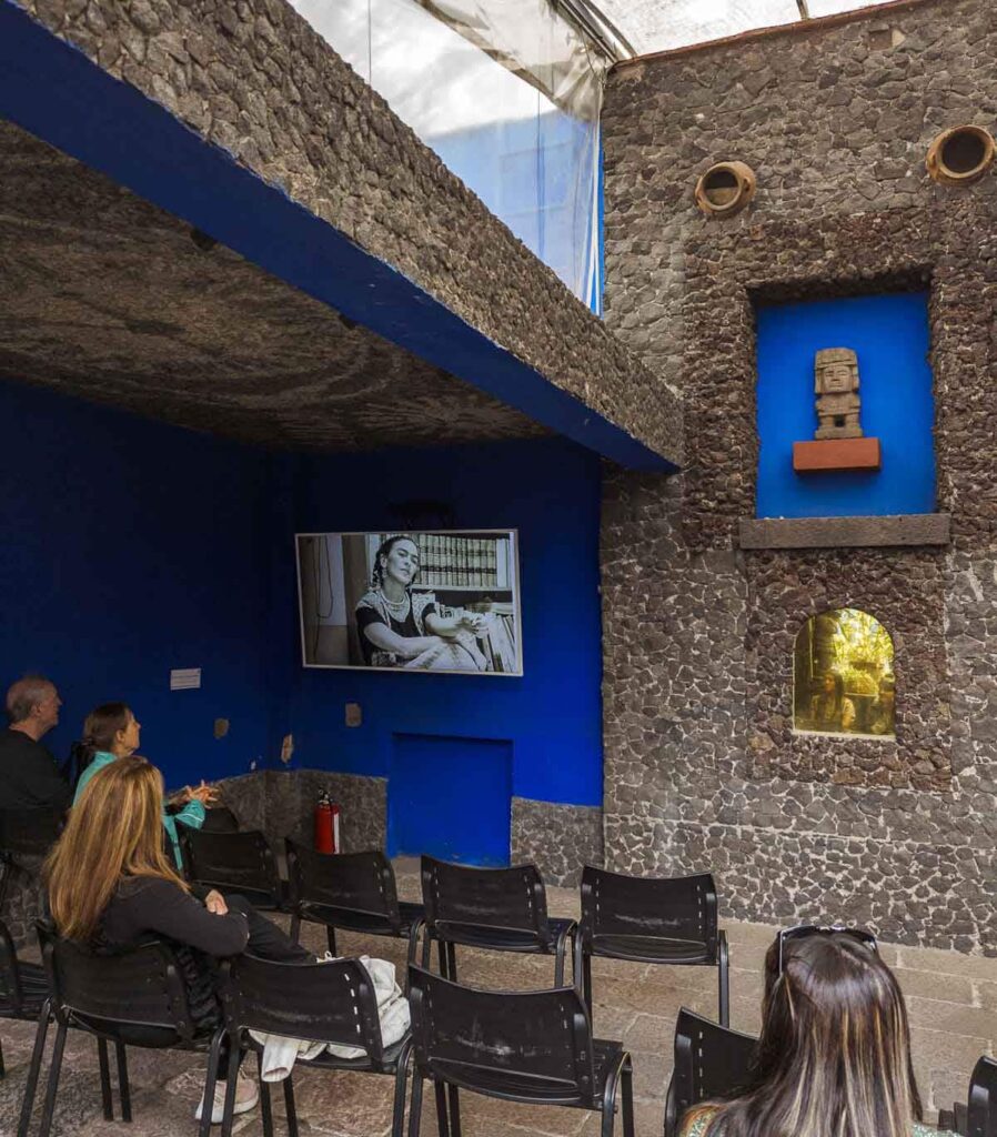 Group of people captivated by Frida Kahlo Museum in Mexico, attentively watching TV display showcasing her remarkable art and life.