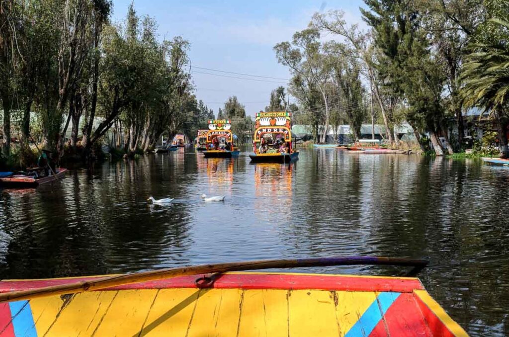 On a colorful yellow, blue and red trajinero looking towards other colorful boats called Daniel and Andre in the middle of the canal at Xochimilco.