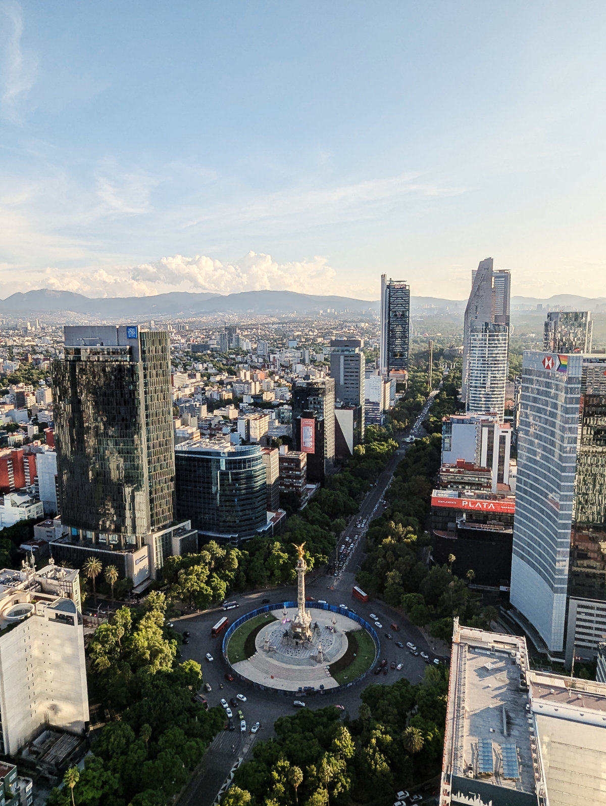 A view of the Angel of Independence from Cityzen rooftop in Mexico City. Buses and cars drive around the roundabout below the angel while sky scrapers line the tree-lined avenue with mountains in the background.