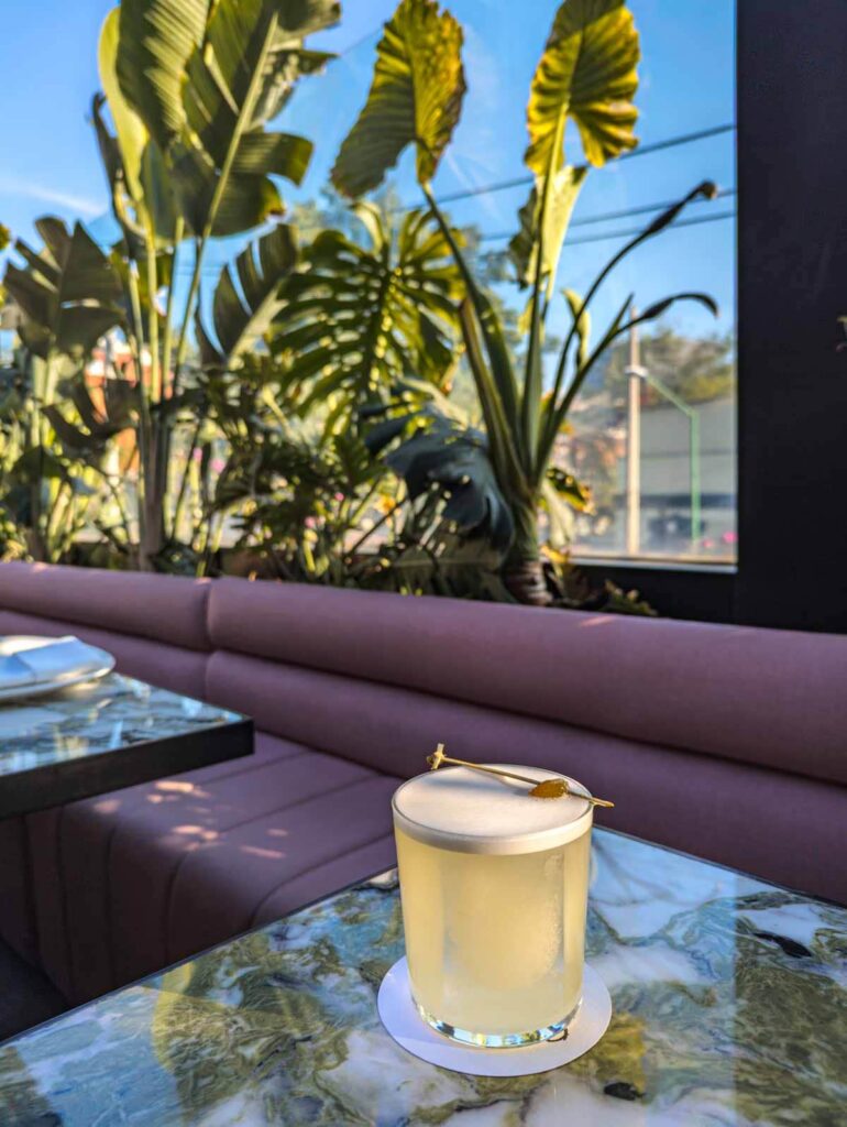 A frothy cocktail on a table at Balmori rooftop bar in Mexico City. Behind the cocktail is a pink couch and tropical plants.