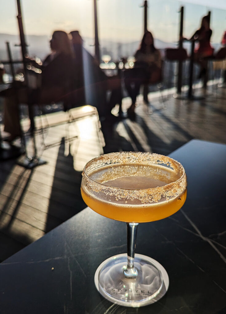 A yellow cocktail with sugar rim at Cityzen Sofitel rooftop in Mexico City is backlit by the setting sun. In the background, blurred people sit at tables along the edge of the rooftop.