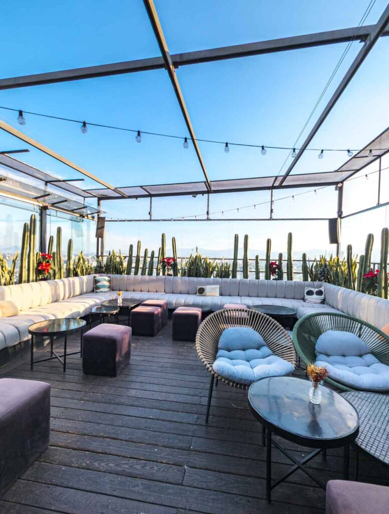 The lounge at Supra Rooftop in Mexico City features long grey couches and other cushioned seating surrounded by tall skinny organo cactus and views of CDMX.