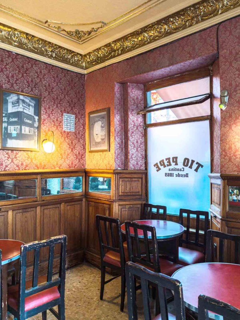 Small red round tables and wooden chairs sit in front of a window with the words Tio Pepe Cantina Desde 1869. Inside the walls are covered in red wall paper with images from the Mexico City cantina's past.