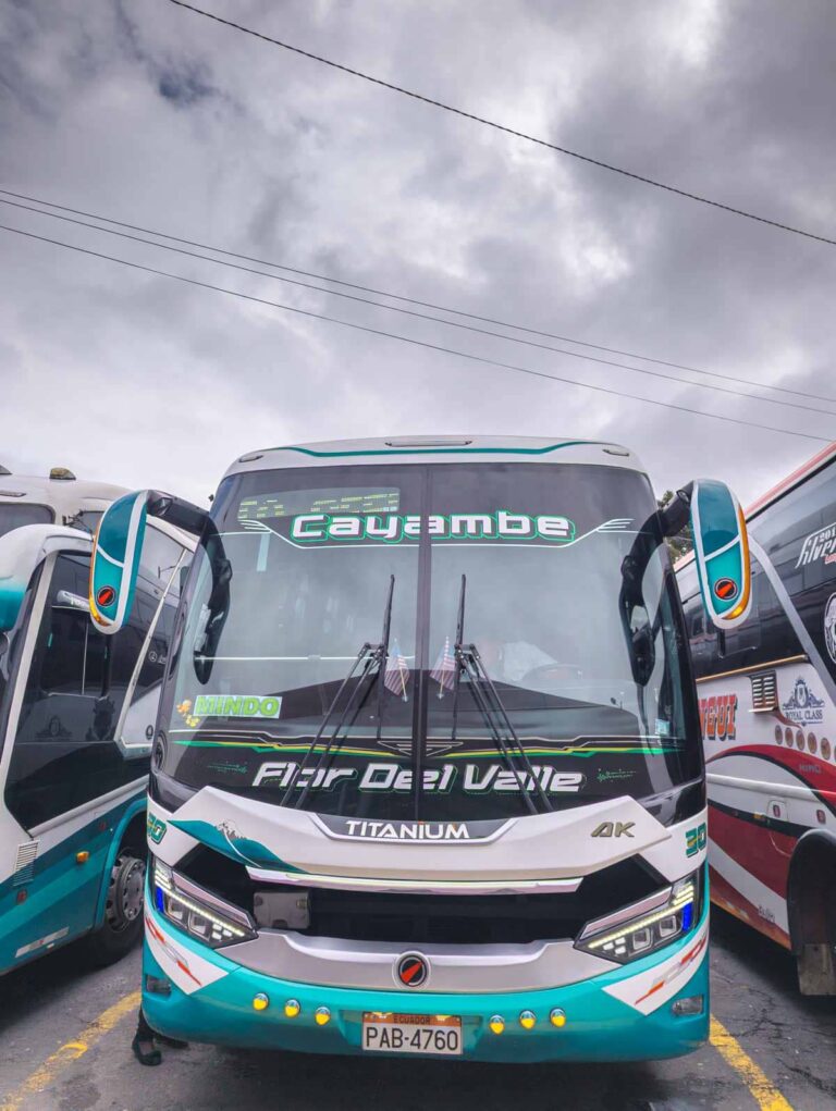 A bus from Quito to Mindo Ecuador detailed with turquoise, black, and grey is parked with a green sign in the windshield that says Mindo.