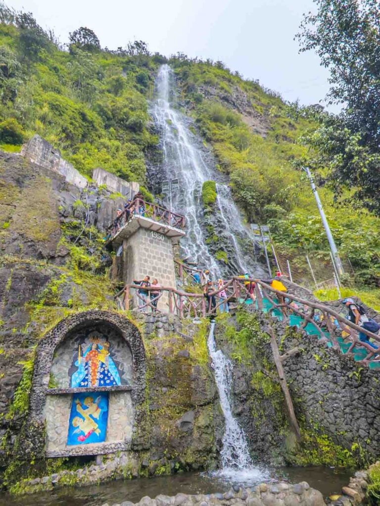 A thin waterfall flows down a steep rocky cliff at Termas de la Virgen in Baños Ecuador, with visitors climbing stairs alongside it. Colorful religious mosaics are built into the stone at the bottom.