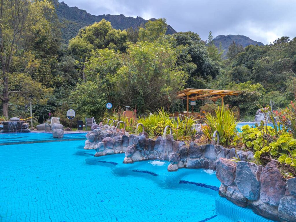 Large hot spring at Papallacta near Quito with a rock wall lined with plants and piped water massage jets. In the background you can see the mountain tops.