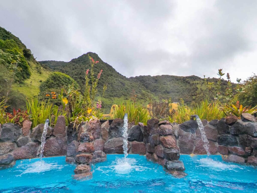 A close up view of three piped water jets coming out of a rock wall at the Papallacta Hot Springs near Quito. The wall is lined with plants and lush green mountains are in the background.